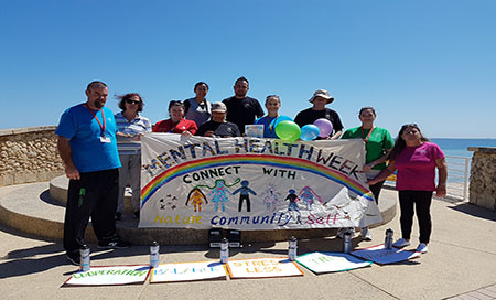 Group of people at beach holding colourful banner reading 'Mental Health Week'